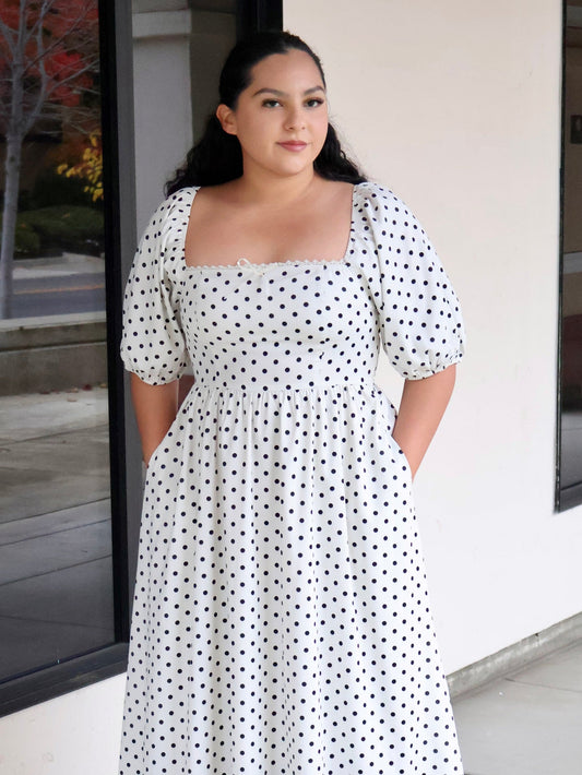Woman wearing a white dress with black polka dots standing outside a building.