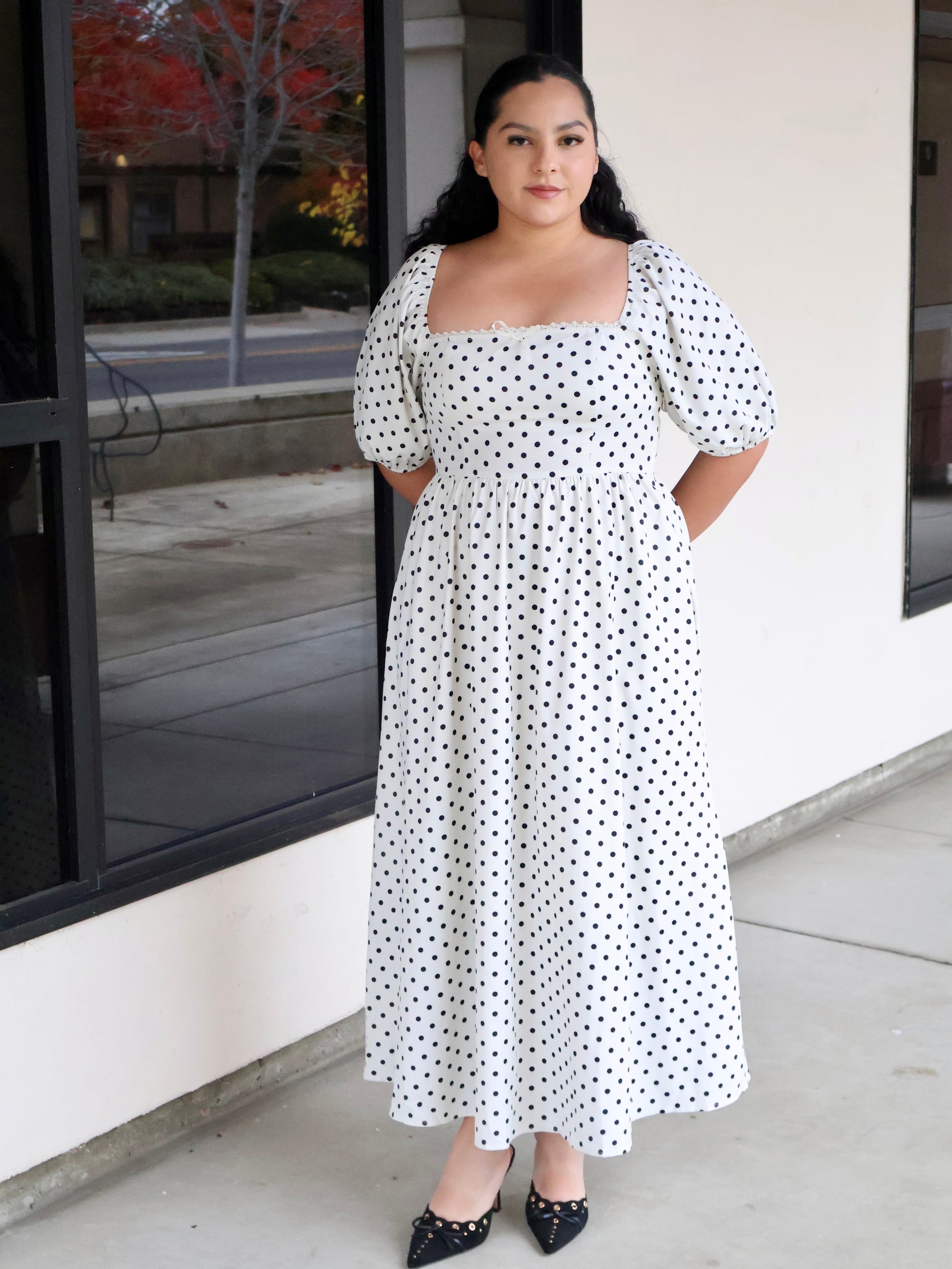 Woman wearing a white dress with black polka dots standing outside a building.